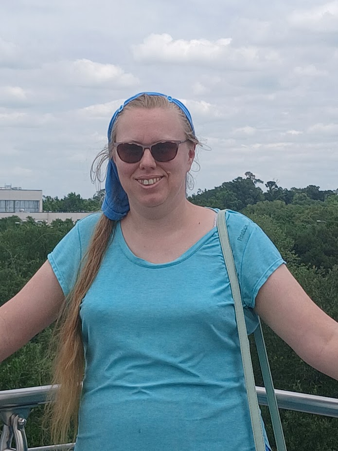 Smiling person wearing sunglasses and a blue headband with a long ponytail, standing outdoors with trees and buildings in the background